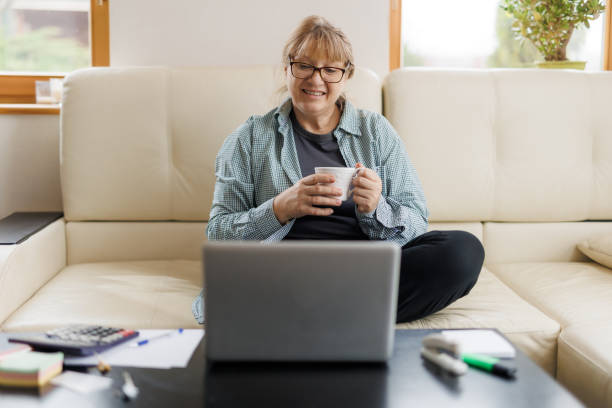 istock Serene mature female with blond hair having hot tea or coffee while working remotely in front of laptop at home 1440761791