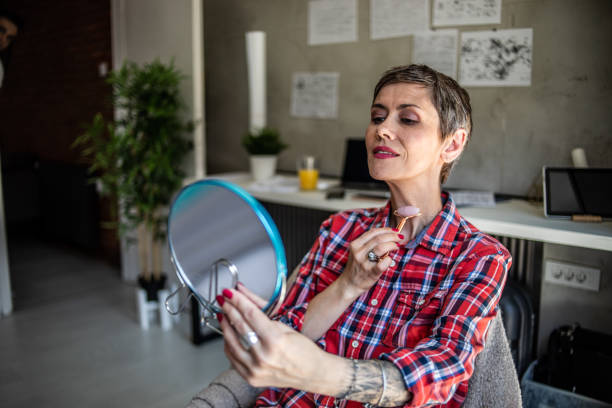 a mature woman with short hair is massaging her face with a derma roller while sitting in her workspace in the apartment - looking glass rock imagens e fotografias de stock