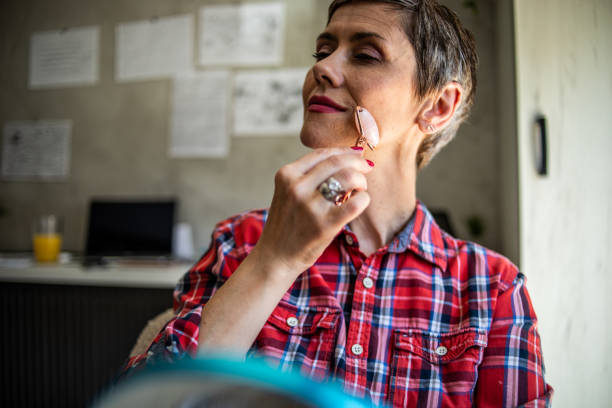 a mature woman with short hair is massaging her face with a derma roller while sitting in her workspace in the apartment - looking glass rock imagens e fotografias de stock