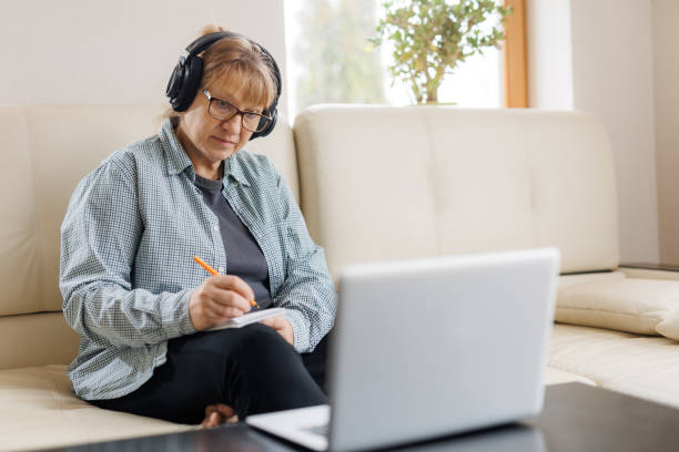 istock Modern middle aged blond woman in headphones take online course or training on computer at home. Smart mature Caucasian female in earphones watch webinar make note study distant on laptop. 1440762677