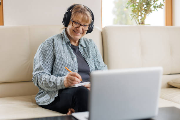 istock Modern middle aged blond woman in headphones take online course or training on computer at home. Smart mature Caucasian female in earphones watch webinar make note study distant on laptop. 1440762709