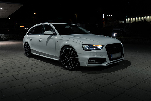 Ulricehamn, Sweden – August 04, 2021: The front part of white Audi A4 Avant with lit headlights outdoors at night