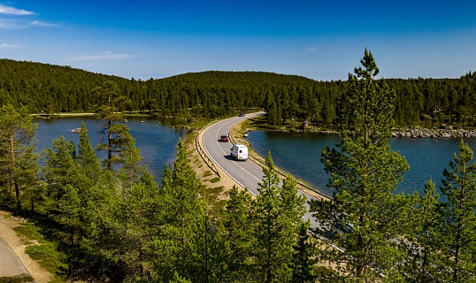 Aerial view of motorhome and cars driving on a asphalt road in middle of forest, lake and tunturi mountains, in Inari, Lapland, Finland