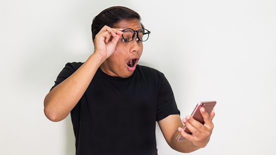 An Asian male with a shocked facial expression in a black t-shirt with glasses looking at his smartphone