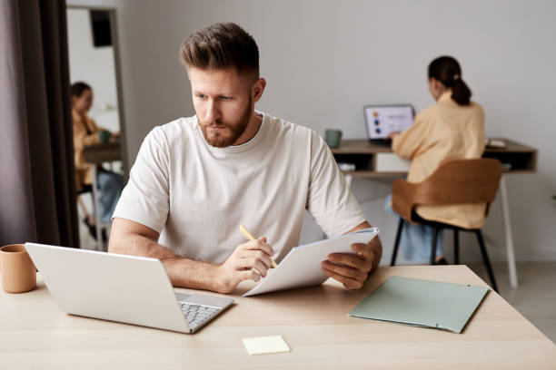 istock Young serious man making notes in copybook and looking at laptop screen 1440777387