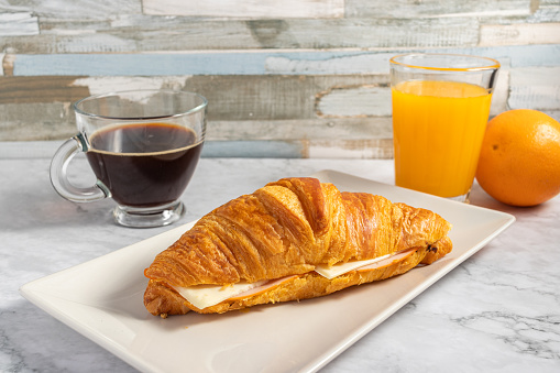 A crunchy delicious croissant with ham and cheese filling, a cup of hot coffee and juice on a table