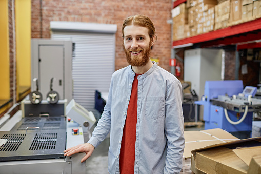 Waist up portrait of smiling young man working in industrial printing shop and looking at camera, copy space