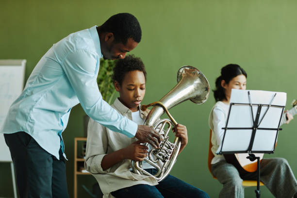 Young confident African American teacher of music consulting schoolboy Young confident African American teacher of music consulting diligent schoolboy with trumpet against pre-teen girl playing guitar brass instrument stock pictures, royalty-free photos & images