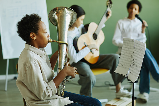 Side view of pre-teen schoolboy blowing wind instrument against two intercultural classmates playing guitar and singing in microphone