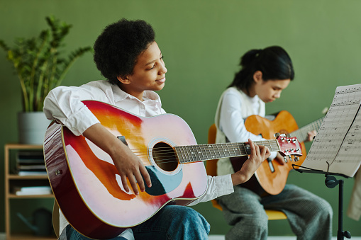 Diligent African American schoolgirl with acoustic guitar looking at paper with notes on music stand while sitting against his classmate