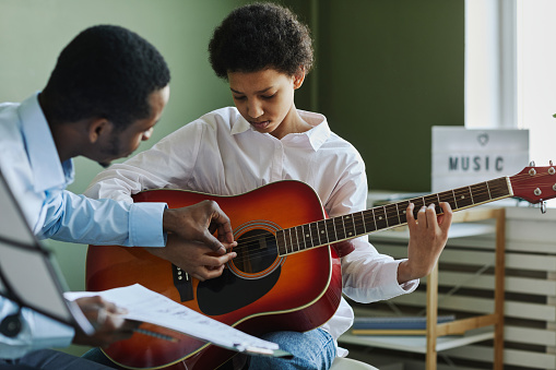 Youthful diligent girl playing acoustic guitar while sitting in front of African American male teacher consulting her at lesson