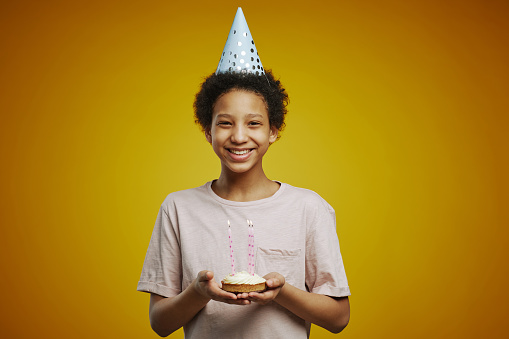 Cheerful African American pre-teen girl with piece of birthday cake with several burning candles standing in front of camera in isolation