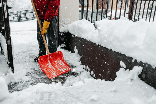 Man in red jacket with snow shovel cleans sidewalks and city street in winter. Cloudy winter day.