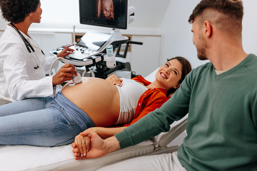 A happy Caucasian couple are at an ultrasound check up at the maternity clinic. The husband is holding his wife's hand as she's lying on the bed.