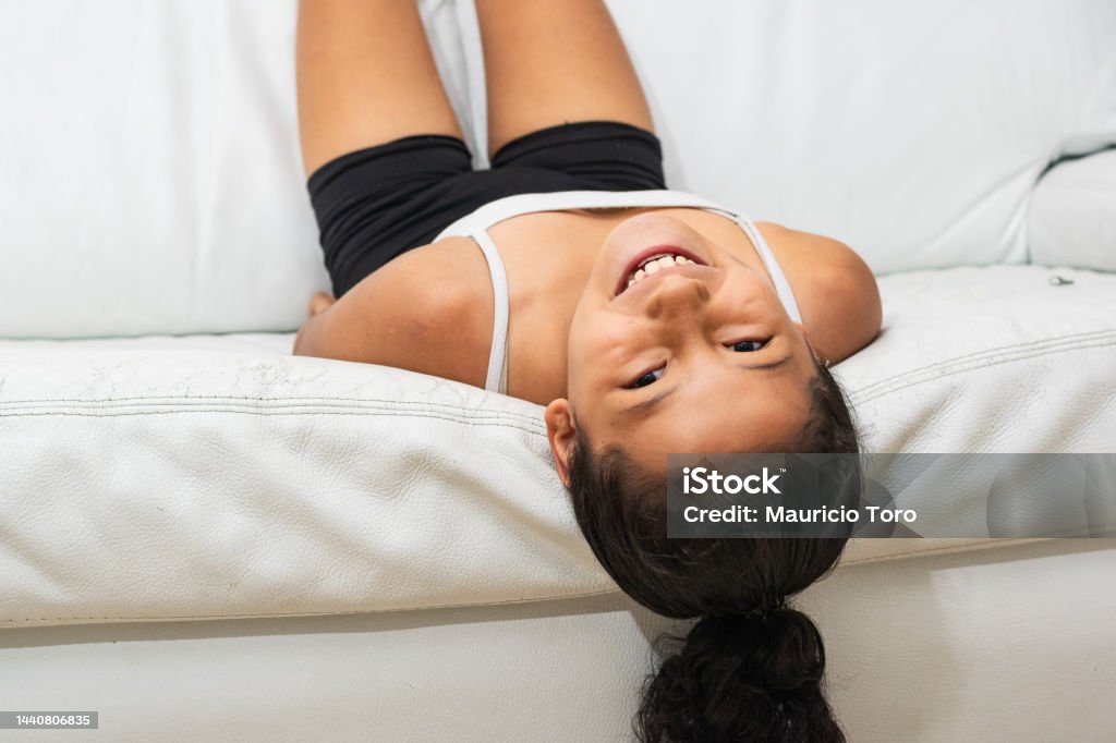 portrait of brunette latina girl, lying on her back on the living room sofa playing and having fun, she has a nice happy face and a big smile. Brown Hair Stock Photo