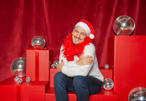 Young handsome smiling man wearing Santa hat and boa sitting on the red background with shining disco balls. New Year style