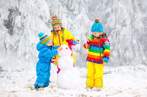 Children build snowman. Kids building snow man playing outdoors on sunny snowy winter day. Outdoor family fun on Christmas vacation. Boy and girl play snow balls. Winter clothing for baby and toddler.