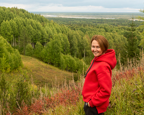 a smiling woman with red hair and a red hoodie on a wooded hill against the background of a forest and a winding river in the distance. Arkhangelsk region Pinezhsky district , Golubino Nature Reserve Park