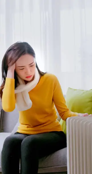 asian woman wearing scarf feel cold and the heating is broken in winter sitting on couch at home