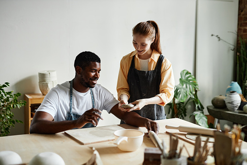 Warm toned portrait of smiling multirational couple enjoying art class in pottery studio together, copy space