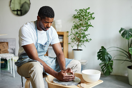 Portrait of black young man using pottery wheel and creating handmade ceramics in cozy studio with green plants