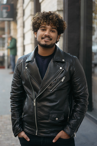 Portrait of smiling handsome Indian fashion model wearing black leather jacket posing for picture on the street. Fashion concept