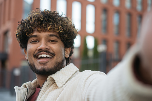 Smiling modern Indian man taking selfie, having video call on the street. Emotional curly haired asian influencer recording video, communication online outdoors. Wireless technology concept