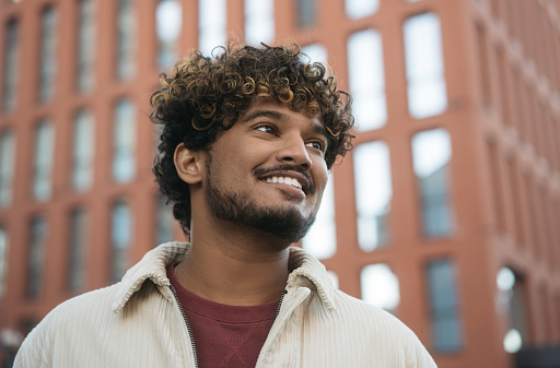 Closeup portrait of smiling Indian man with stylish curly hair looking away on the street. Handsome asian fashion model wearing trendy jacket posing for picture outdoors, copy space