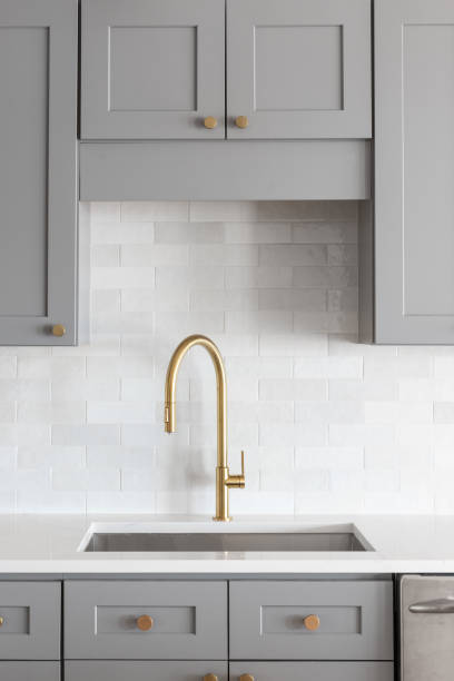 A gold faucet in a grey and white kitchen. A kitchen sink detail shot with a gold faucet, marble backsplash, grey cabinets, and gold hardware. cleaning cupboard stock pictures, royalty-free photos & images