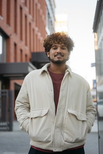 Smiling handsome Indian man wearing stylish casual jacket looking at camera on the street. Happy asian fashion model with curly hair posing for picture outdoors