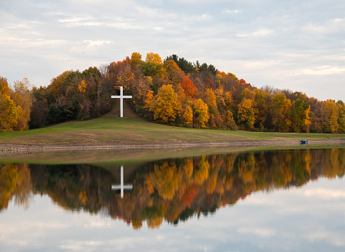 Photograph of a white Cross at the edge of the forest in autumn, the leaves are changing and the cross is reflected in the water.