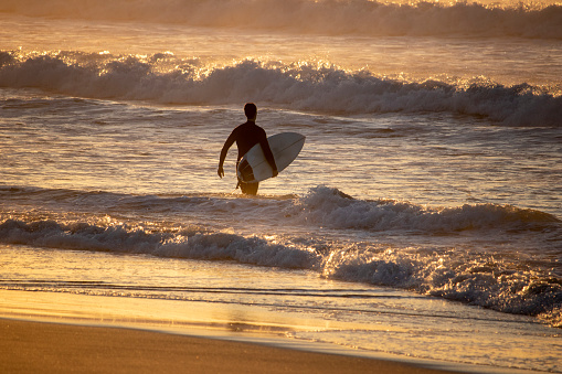 A surfer is walking along the beach with his surf board and is entering the surfA surfer is walking on the sand along a beach with his surf board and is observing the surf on a beach at sunrise
