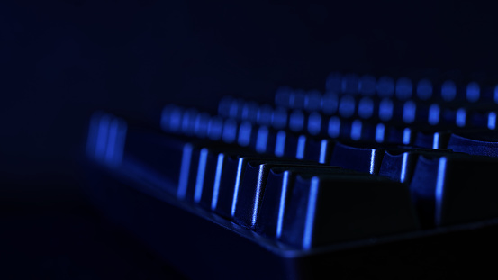 Black mechanical gaming computer keyboard illuminated in the dark by blue light from a monitor screen. Side view. Copy space. Macro. Selective focus. Depth of space and bokeh. Close-up