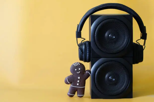 Photo of Two black audio speakers, wireless headphones and cheerful gingerbread man on a yellow background. Concept of a holiday party, disco, pop music, family and children's audio broadcasts.