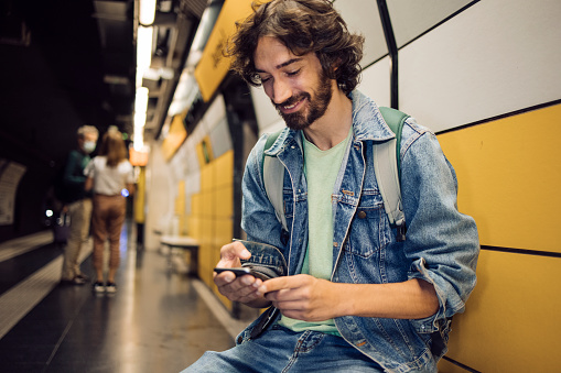 Portrait of a young man on vacation in Barcelona waiting for his train in the subway and using his smartphone.