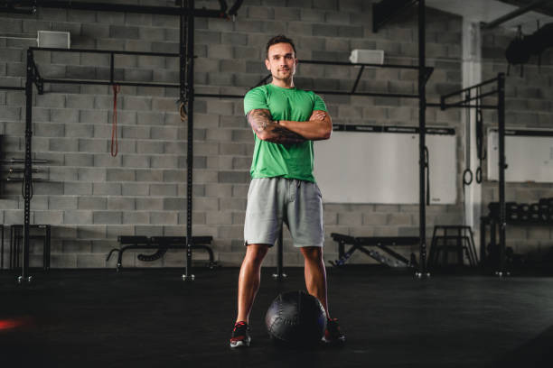 Gym Portrait of Confident Young Man with Medicine Ball Full length front view of Caucasian man in late 20s with arms crossed and looking at camera while standing over medicine ball. running shorts stock pictures, royalty-free photos & images