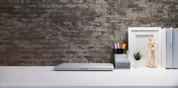 istock Laptop computer, picture frame, books and pencil holder on white table against brick wall. 1440868491