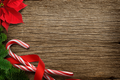 A Christmas background of candy canes wrapped in a red ribbon and a red poinsettia framed by a bough from a Christmas tree on a richly textured wood surface that provides ample room for copy and text.