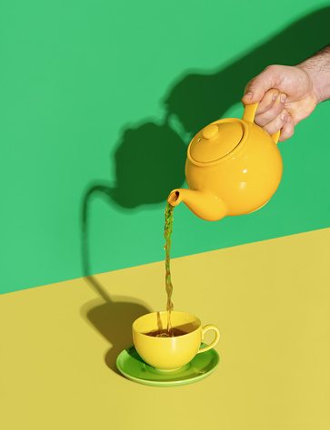 Pouring hot mint tea in a cup, in bright light on a yellow colored table. Pouring tea from a yellow teapot.