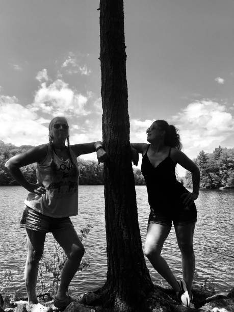 Active senior mom and adult daughter stopping on an island during a pontoon adventure on The Menominee River in Wisconsin, USA. The playful pair are having fun and both are leaning against a big tree in the middle of the photo. Both are active and adventurous, with similar body types and big smiles, enjoying the nature outing. The Menominee River can be seen in the background. Photo in black and white.
