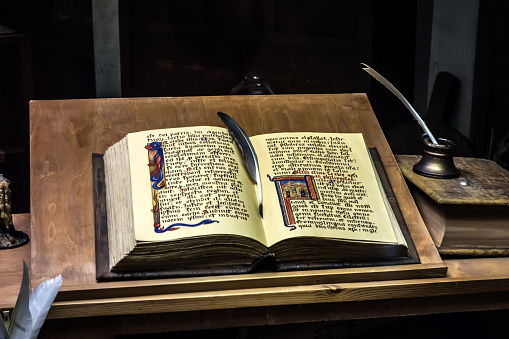 Open ancient handwritten book with pictures lies on a wooden table. Near the book inkwell with quill pen. Vintage still life.