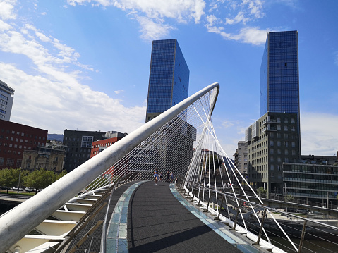 Tourists walking on the futuristic Zubizuri pedestrian bridge in Bilbao, Spain. In the background, the Isozaki Atea twin towers, the tallest residential buildings in the city and the Basque Country, 83 metres tall.