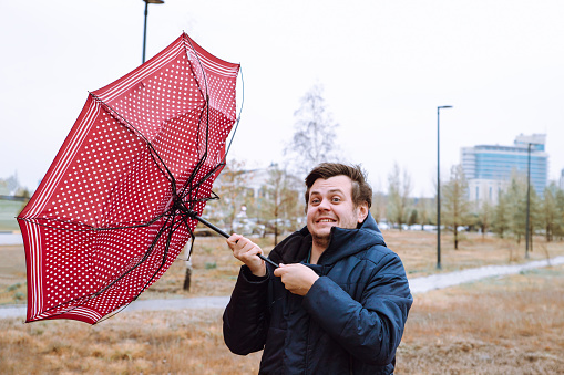 Un joven blanco europeo conmocionado tontea con sombrillas en el parque de la ciudad, fuertes vientos de tormenta, lluvia y mal tiempo. El viento rompió el paraguas. Retrato de estilo de vida de cintura hacia arriba de otoño. photo