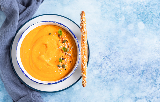 Bright vegetarian autumn pumpkin and carrot soup with seeds, mint and spices, blue concrete background. Comfort food, fall and winter healthy slow food concept.