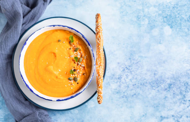 Bright vegetarian autumn pumpkin and carrot soup with seeds, mint and spices, blue background. Comfort food, fall and winter healthy slow food concept. Bright vegetarian autumn pumpkin and carrot soup with seeds, mint and spices, blue concrete background. Comfort food, fall and winter healthy slow food concept. squash soup stock pictures, royalty-free photos & images