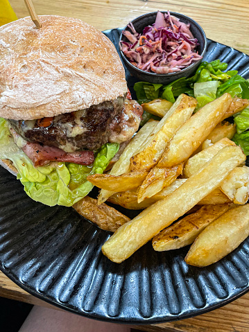Photo showing a black plate with hamburger / cheese burger, which is served with chunky chips with potato skins / French fries, lettuce, bacon, floured burger bap bread roll, salad and homemade coleslaw, with carrot, onion, mayonnaise and red cabbage