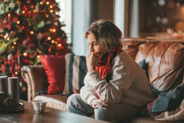 Young teen boy with long hair thoughtful look sad eyes negative mood angry and crying at home. Stylish zoomer gen Z pensive on new year holidays with xmas tree bokeh lights garlands eve 25 december