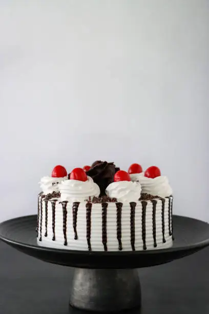 Stock photo showing an elevated view of a homemade, luxury, Black Forest gateau on a black cake stand, a chocolate cake decorated with white icing, morello cherries, whipped cream, melted chocolate with chocolate shavings displayed on a black cake stand against a white background.