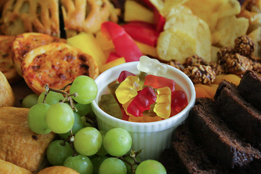 Stock photo showing close-up view of party food buffet with wooden charcuterie board covered with prepared sliced and chopped ingredients to make a combined dessert and savoury grazing board. The display includes sausage rolls, red and white grapes, ramekins of chocolate pieces and gummy bear sweets, red and yellow bell peppers, dried apricots, chocolate covered popcorn, mini quiches, cubes of hard cheese, pork pie, pecans, potato crisps, chocolate cake and bread.