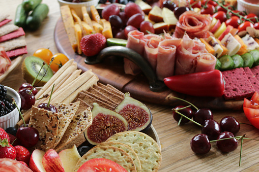 Stock photo showing close-up view of wooden charcuterie board covered with prepared sliced and chopped ingredients including soft and hard cheeses, tomatoes, strawberries, ham and salami roses, rows of crackers, kiwi, vine tomatoes, blueberries, figs, ramekins of honey and chutney, peppers, grapes, olives and cherries.